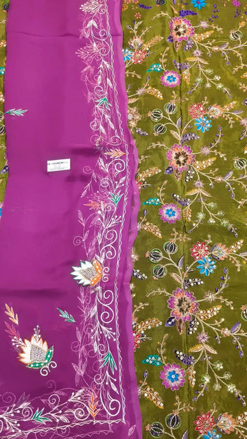 A close-up of a traditional embroidered fabric, likely a saree, featuring a rich magenta purple border with white floral embroidery and a contrasting olive green body adorned with dense multicolor floral embroidery.