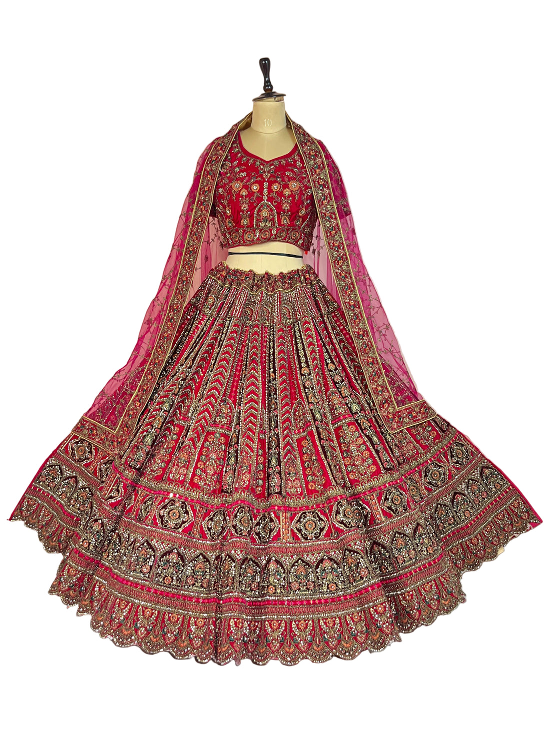 A woman stands in a decorated indoor setting wearing an ornate pink and red embroidered traditional outfit with a matching sheer dupatta, heavily detailed with gold and beige patterns. The scene includes floral arrangements, hanging white garlands, and lit candles.