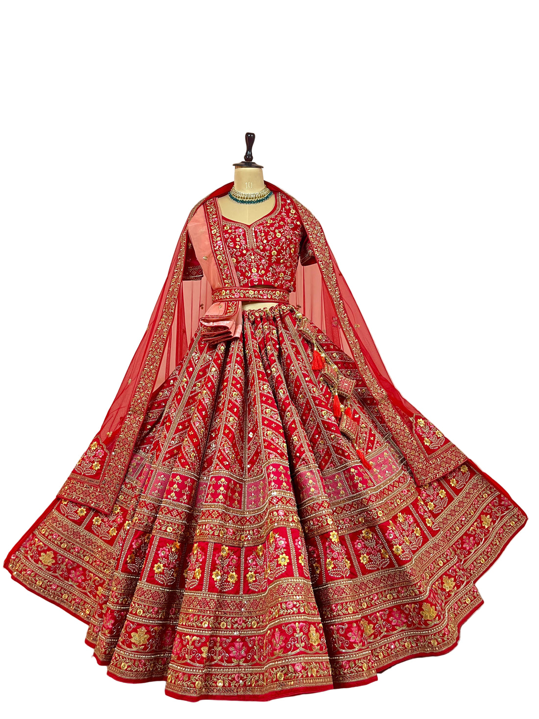 A woman seated on a white cane chair wearing an ornate red and gold traditional bridal lehenga choli set in an indoor room with pampas grass decor.