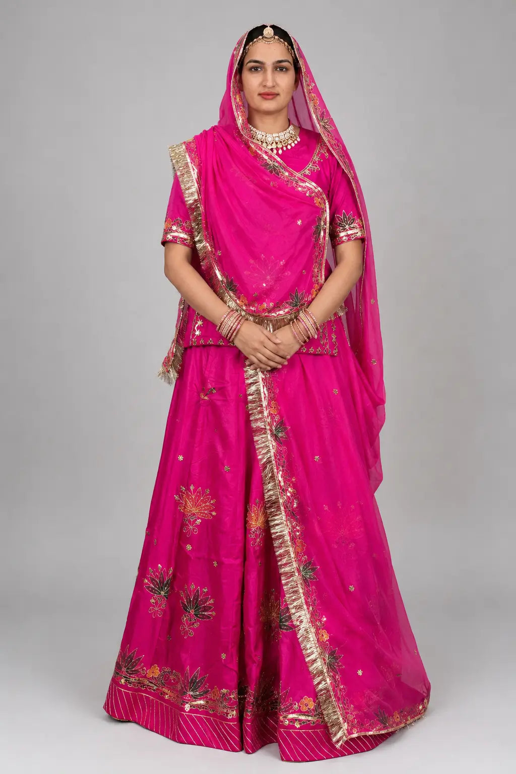 A woman modeling a bright pink traditional Indian outfit with gold embroidery and a matching sheer dupatta, photographed against a plain light gray studio background.