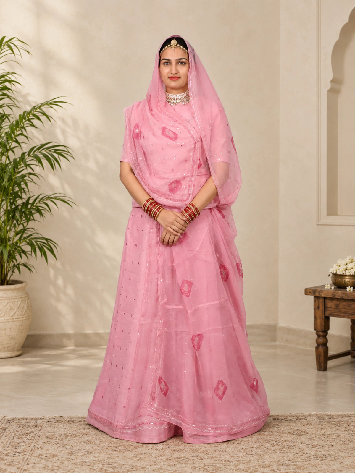 A woman modeling a pink traditional Indian outfit with a matching sheer dupatta, standing in an indoor setting with a beige wall, potted plant, and wooden table.