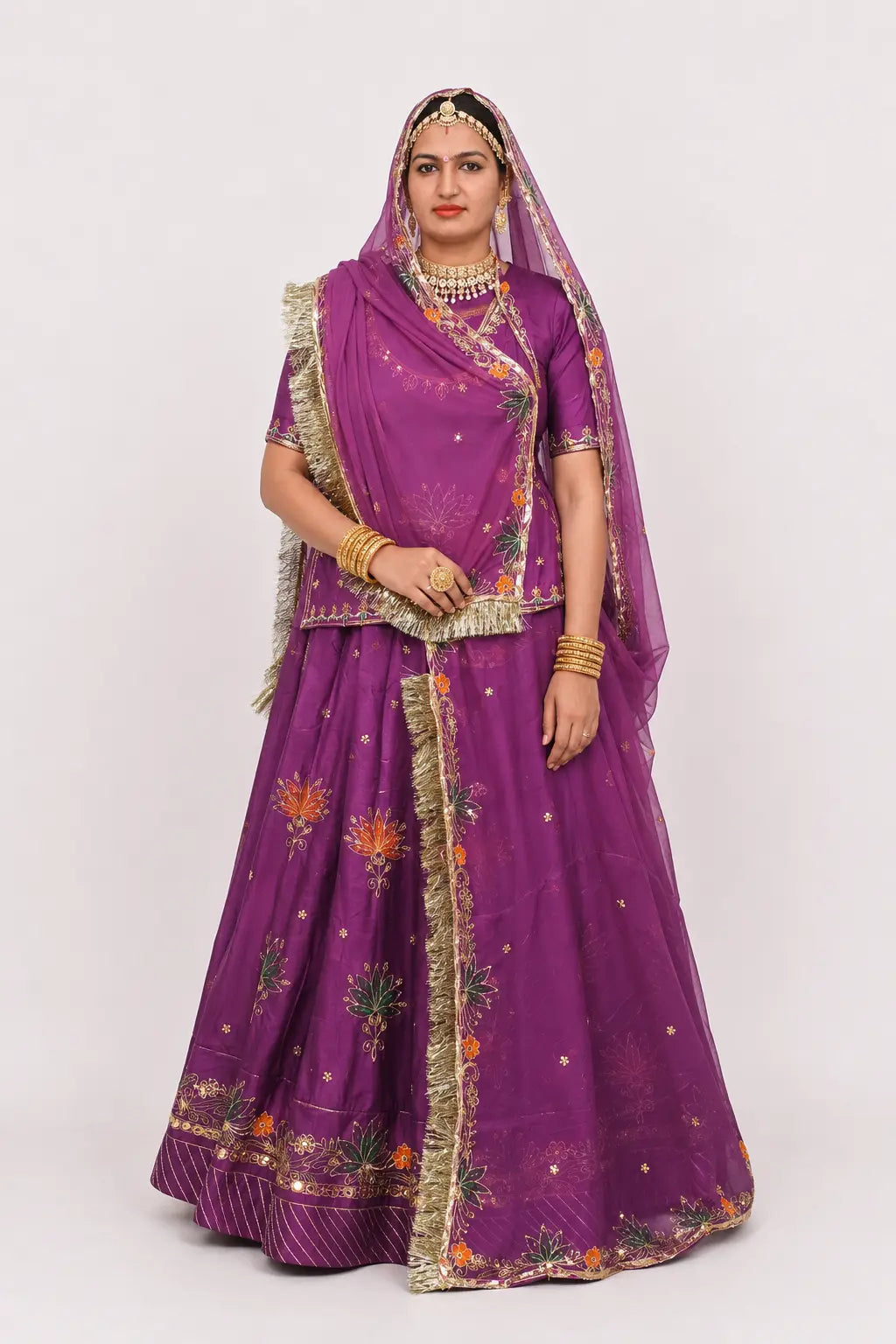 A woman modeling a traditional Indian outfit consisting of a purple lehenga choli set with gold embroidery and a matching sheer dupatta, set against a plain light gray background.