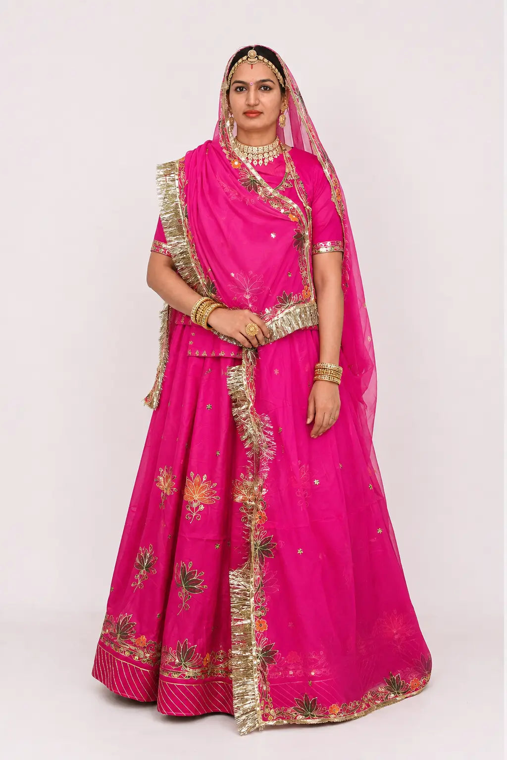 A woman modeling a bright pink traditional Indian outfit with gold embroidery and a matching sheer veil, photographed against a plain light gray studio background.