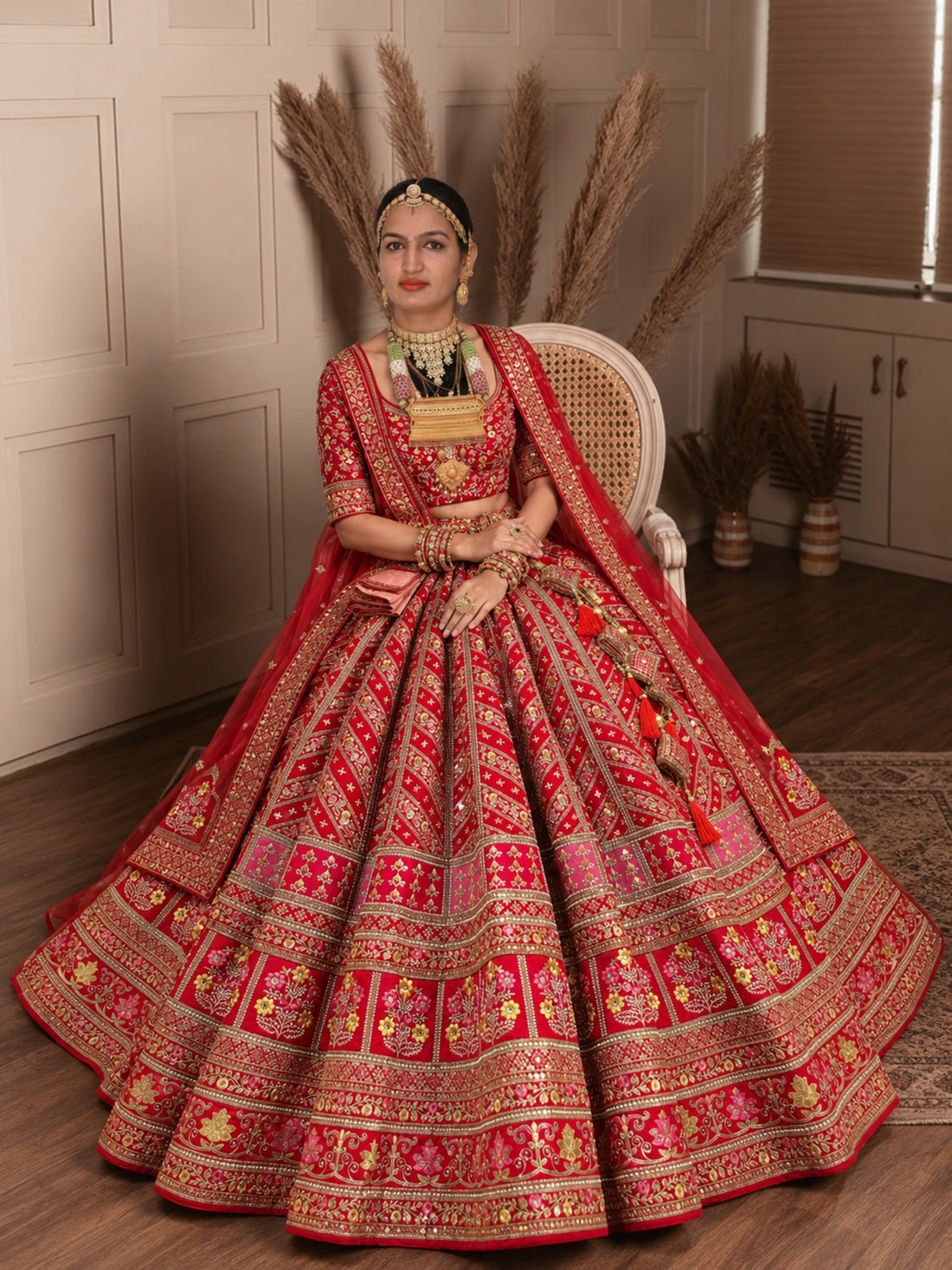 A woman seated on a white cane chair wearing an ornate red and gold traditional bridal lehenga choli set in an indoor room with pampas grass decor.