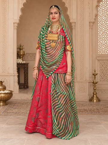A woman models a traditional Indian rajputi suit outfit in sage green and black with gold embroidery, set against an ornate, historic interior with arches and brass lamps.