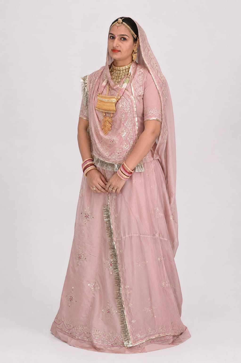 A woman modeling a traditional pink Indian ethnic outfit, featuring a heavily embroidered pink lehenga, matching blouse, and dupatta, styled with gold jewelry against a plain light gray background.