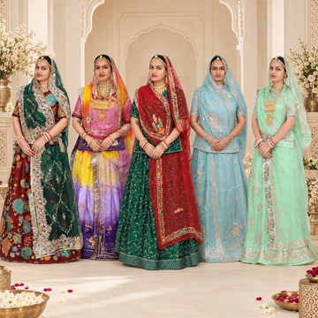 Five women in traditional Indian attire stand together in an ornate indoor setting. They are wearing colorful embroidered lehengas and sarees in green, pink, red, light blue, and mint green, with gold jewelry and veils.