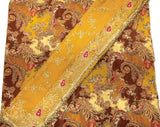 A close-up of a traditional patterned fabric, likely an unstitched salwar suit material, featuring a rich design of brown, mustard yellow, beige, and red paisley motifs with a decorative gold-trimmed border.