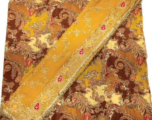A close-up of a traditional patterned fabric, likely an unstitched salwar suit material, featuring a rich design of brown, mustard yellow, beige, and red paisley motifs with a decorative gold-trimmed border.