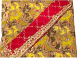 A close-up of a patterned fabric or textile laid flat, featuring a bright red diagonal sash or border with ornate gold embroidery over a yellow and brown paisley background.