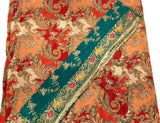 A close-up of a patterned textile, likely a sari or scarf, featuring a diagonal teal border with floral embroidery over a red, orange, and cream paisley background.