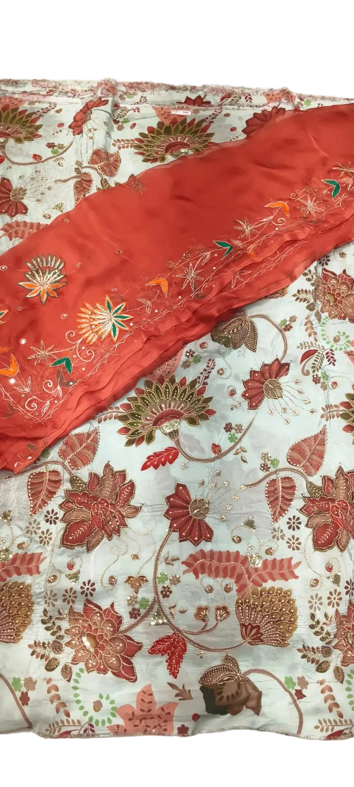 A close-up of a traditional embroidered fabric set, featuring a light blue base with a floral and vine pattern in rust red, gold, and green, accented by a solid rust red border with colorful floral embroidery.