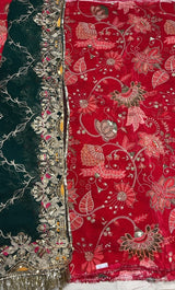 A close-up of a richly embroidered fabric, likely a dupatta or scarf, featuring a deep red side with pink and gold floral motifs and a dark green side with ornate gold embroidery and mirror work.