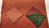 A rust-orange cotton saree fabric with small black and silver mirror work dots, displayed flat with a folded striped brown and orange blouse piece and a folded green and orange dupatta piece on a white background.