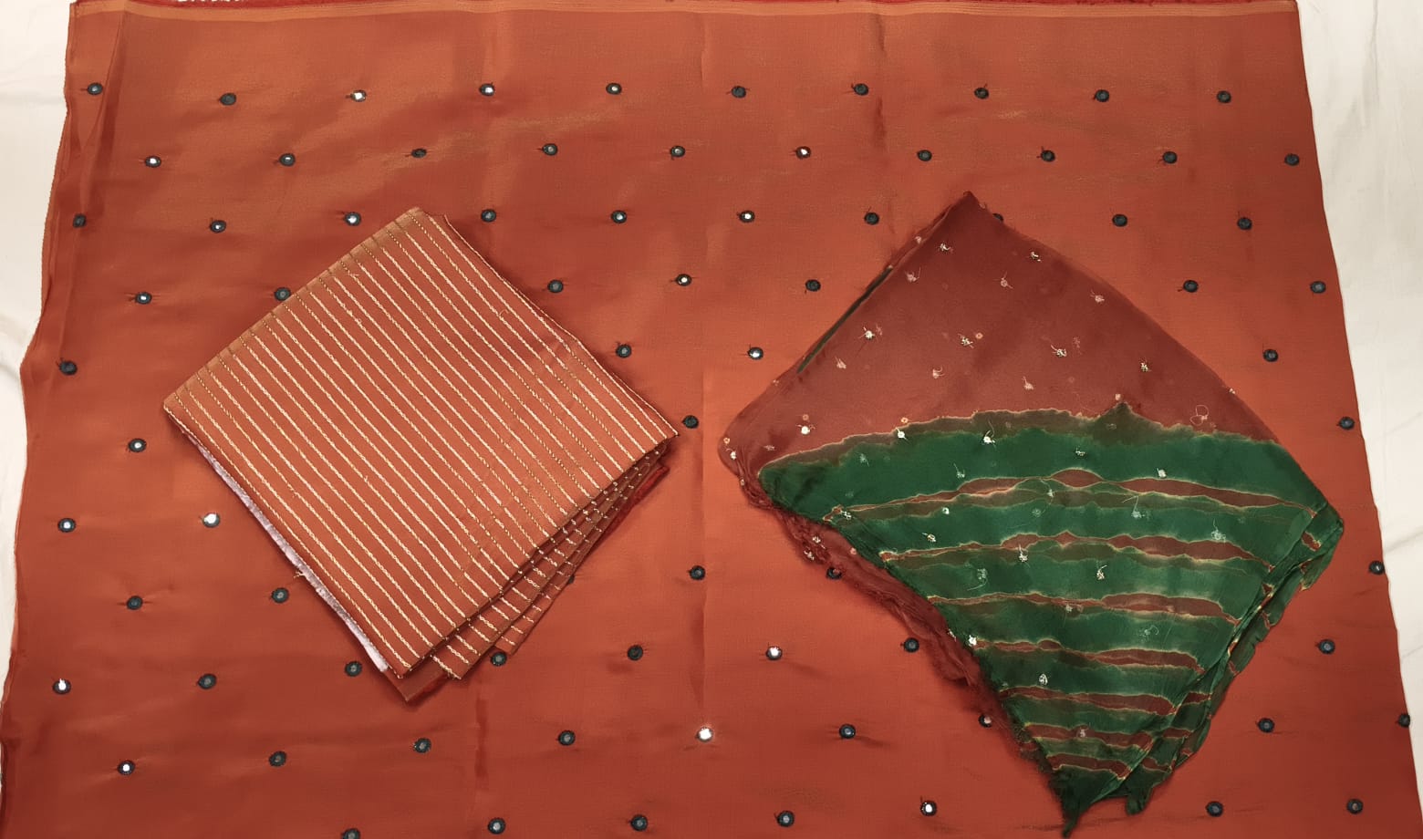 A rust-orange cotton saree fabric with small black and silver mirror work dots, displayed flat with a folded striped brown and orange blouse piece and a folded green and orange dupatta piece on a white background.