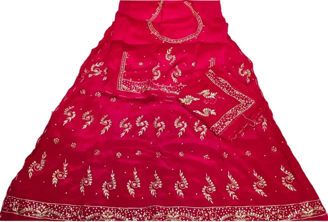 A bright pink traditional Indian dress material set laid flat on a white sheet, featuring gold floral embroidery and mirror work. A small portion of a yellow garment is visible on the right, and parts of a person's legs and hand are visible on the left edge.
