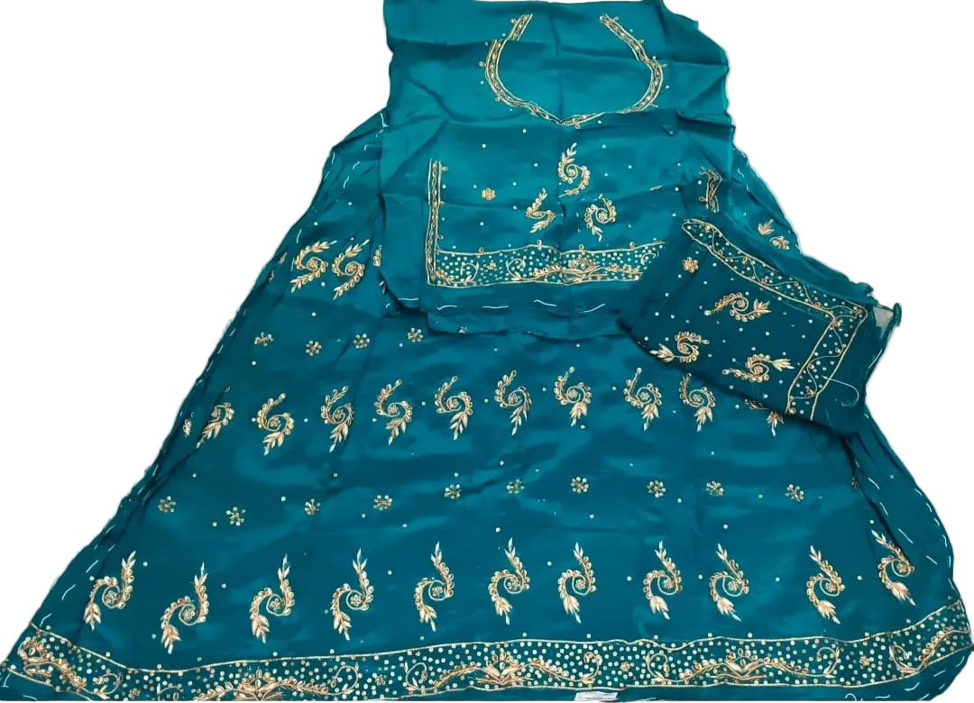 A teal blue traditional outfit laid flat on a light beige fabric background, featuring gold embroidered motifs and borders, with a matching headpiece and dupatta.