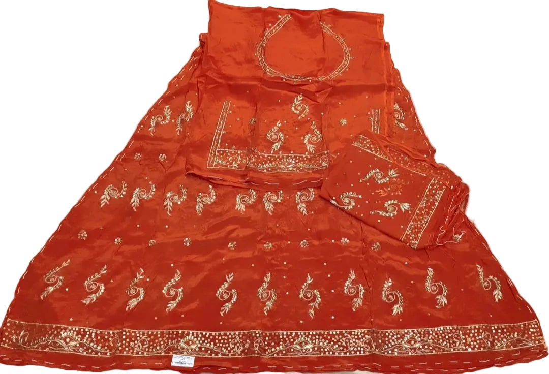 An orange-red embroidered traditional Indian dress set laid flat on a white fabric background. The set includes a flared skirt, a matching sleeveless blouse, and a folded dupatta, decorated with gold and silver mirror work and embroidery.