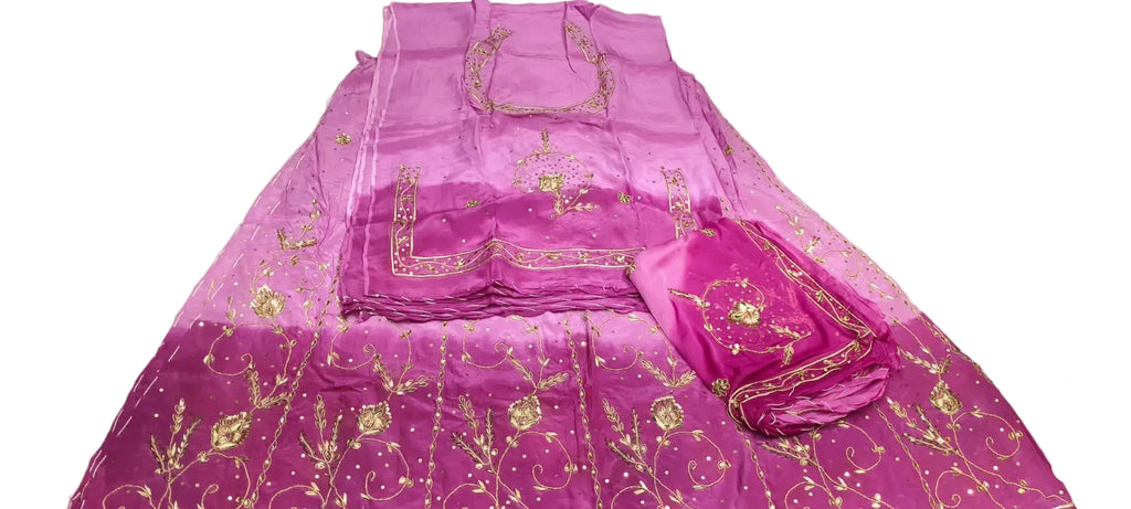 A folded magenta pink embroidered salwar suit set with gold metallic embroidery, displayed on a white floor next to a person's hands and legs.