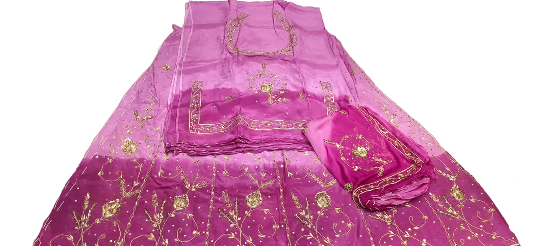 A folded magenta pink embroidered salwar suit set with gold metallic embroidery, displayed on a white floor next to a person's hands and legs.