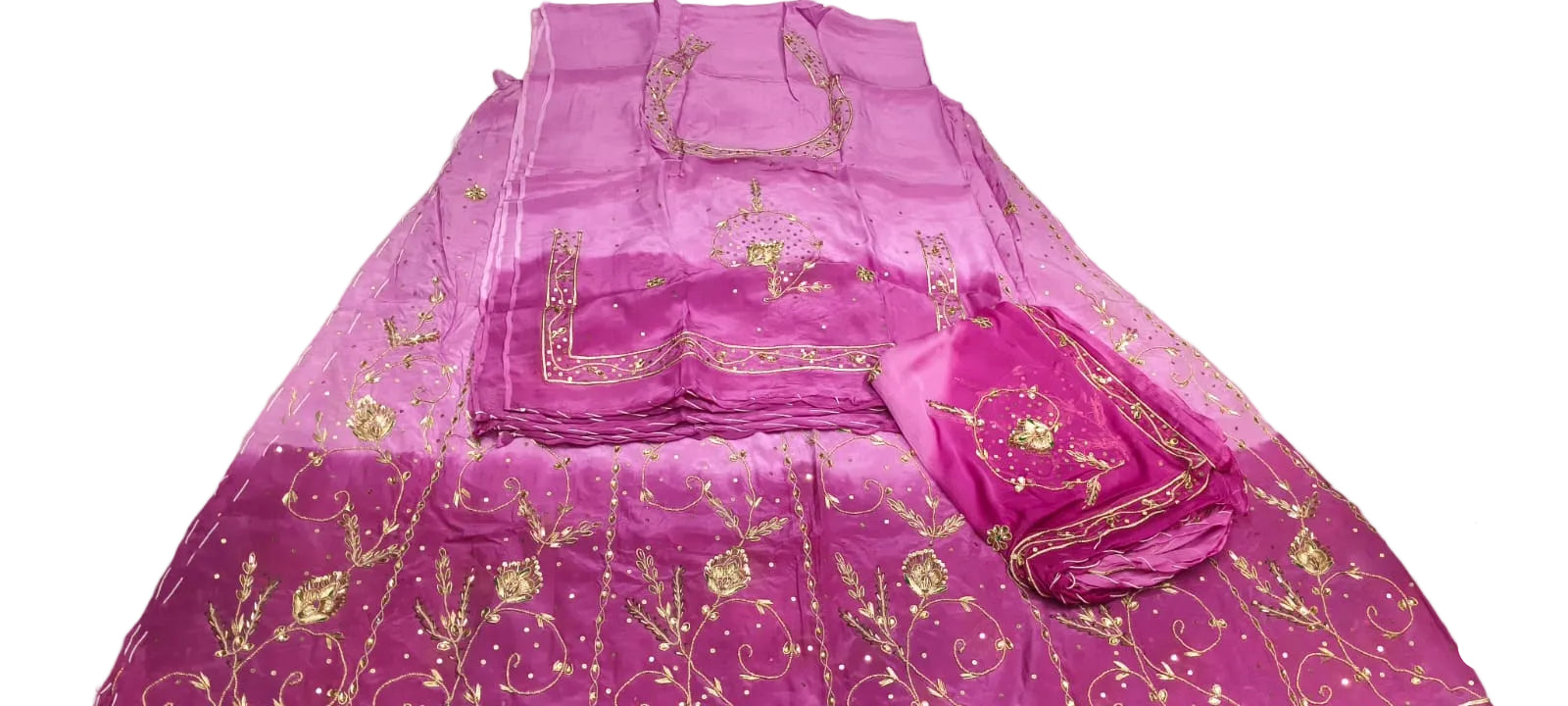 A folded magenta pink embroidered salwar suit set with gold metallic embroidery, displayed on a white floor next to a person's hands and legs.