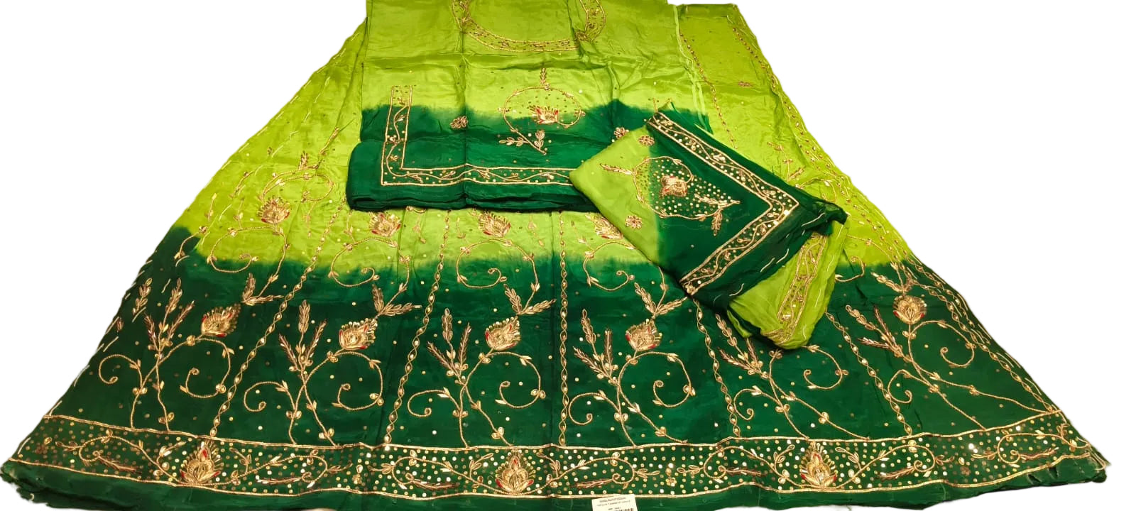 A green and dark green embroidered ethnic skirt set with a matching blouse piece, laid out on a light floor. The skirt features a bright lime green upper section that fades into a dark emerald green hem with intricate gold and silver embroidery.