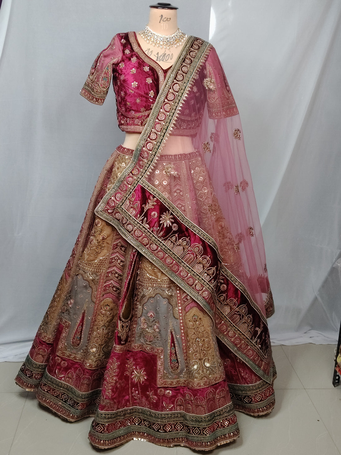 A woman seated in a studio wearing an ornate pink and gold embroidered traditional outfit with a matching sheer dupatta, against a dark burgundy backdrop.