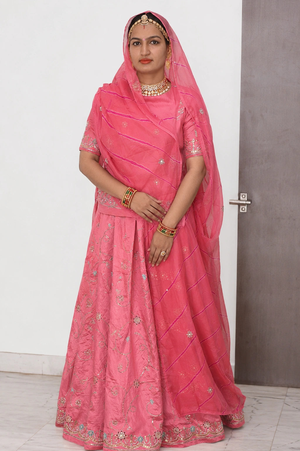 A woman wearing a bright pink traditional Indian outfit with a matching sheer dupatta, embellished with silver mirror work and floral embroidery, standing indoors against a white wall and gray door.
