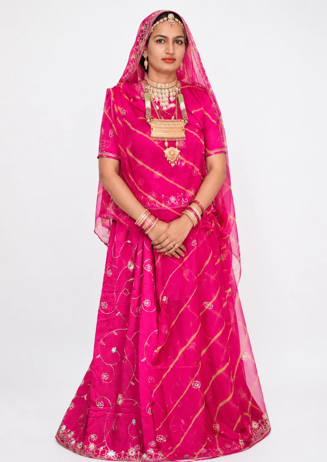 A woman modeling a bright pink traditional Indian dress with gold tie-dye stripes, a matching sheer veil, and ornate gold jewelry against a plain white background.