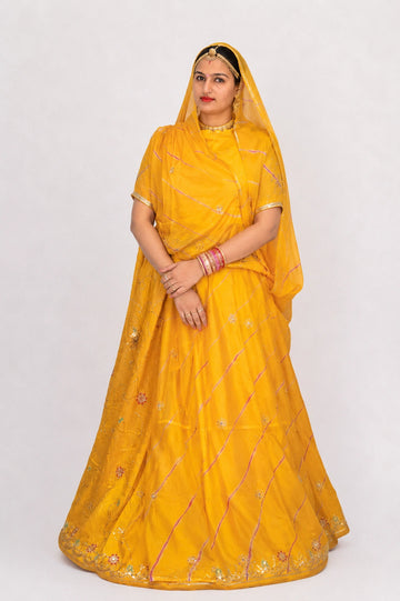 A woman modeling a bright yellow traditional Indian outfit with a matching sheer dupatta, featuring embroidered details and gold jewelry, set against a plain light gray background.