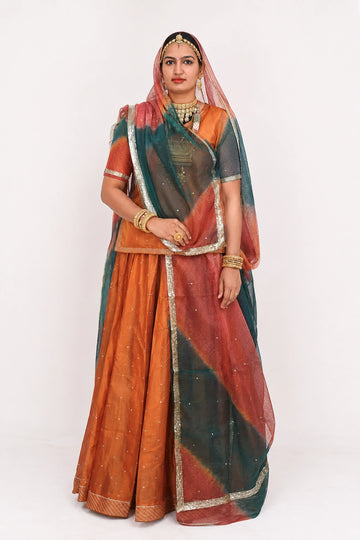 A woman modeling a traditional Indian outfit consisting of an orange skirt, a matching orange blouse with a dark teal sleeve, and a sheer dark teal dupatta with orange and silver borders, set against a plain white studio background.
