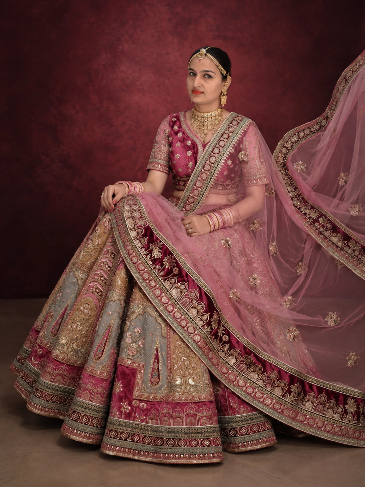A woman seated in a studio wearing an ornate pink and gold embroidered traditional outfit with a matching sheer dupatta, against a dark burgundy backdrop.