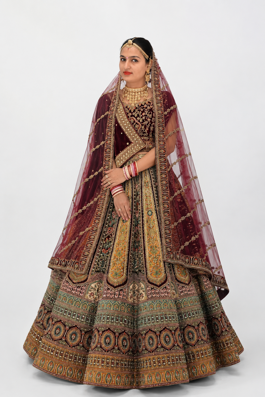 A woman seated on a white chair in front of pink-lit stairs, wearing an ornate traditional bridal outfit with a heavily embroidered voluminous skirt, matching sleeves, and a draped dupatta. She is styled with extensive gold jewelry.