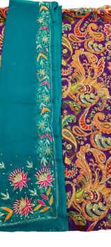 A close-up of a traditional embroidered fabric, showing a teal blue border with pink and orange floral embroidery next to a purple background with an intricate paisley pattern in orange, gold, and green.