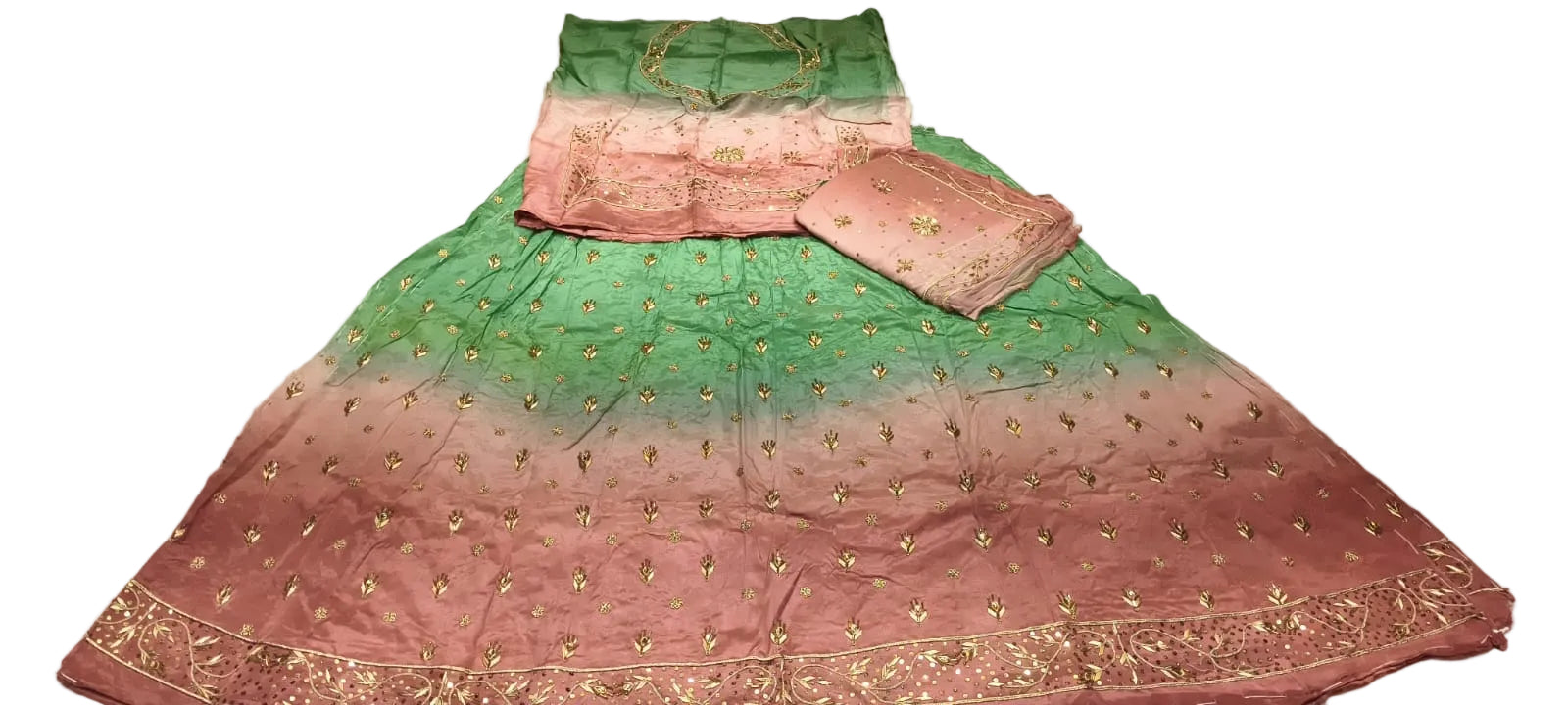 A green and pink gradient embroidered skirt set with matching dupatta and blouse is laid out on a white sheet. The skirt features gold embroidery and a gradient transition from green at the top to pink at the bottom.