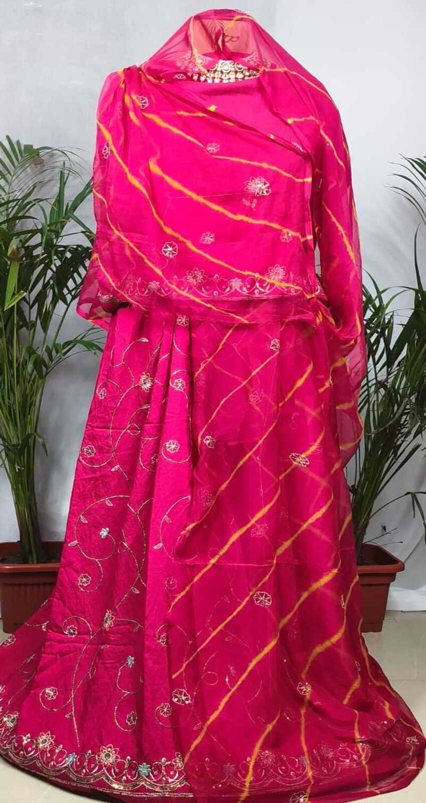 A bright pink traditional outfit displayed on a mannequin, featuring a draped top with a matching sheer dupatta, decorated with gold tie-dye stripes and floral embroidery, set against a background with potted plants.