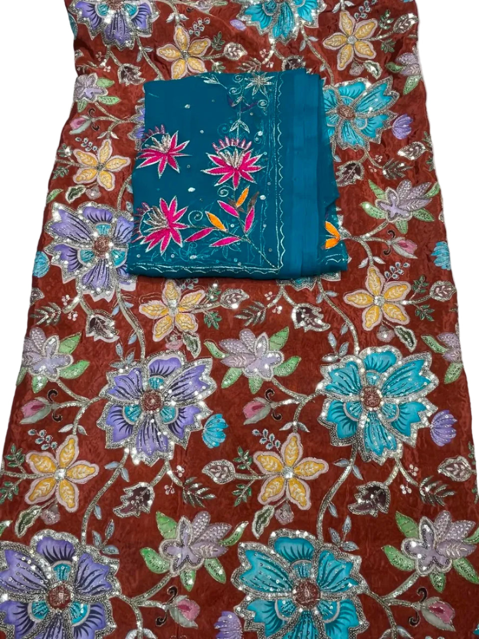 A close-up of a heavily embroidered fabric garment or textile laid flat, showing a rust-brown base with large floral motifs in turquoise, lavender, yellow, and pink, plus metallic silver embroidery and a folded teal fabric piece with floral embroidery placed in the lower center.