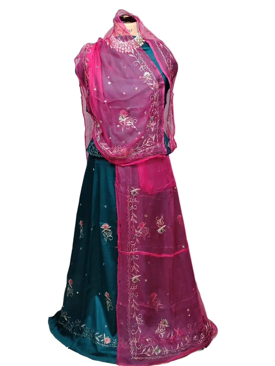 A traditional Indian outfit displayed on a mannequin, featuring a dark teal skirt with floral embroidery, a bright pink sleeveless embroidered overlay, and a matching pink dupatta draped over the shoulders, set against a white fabric backdrop.