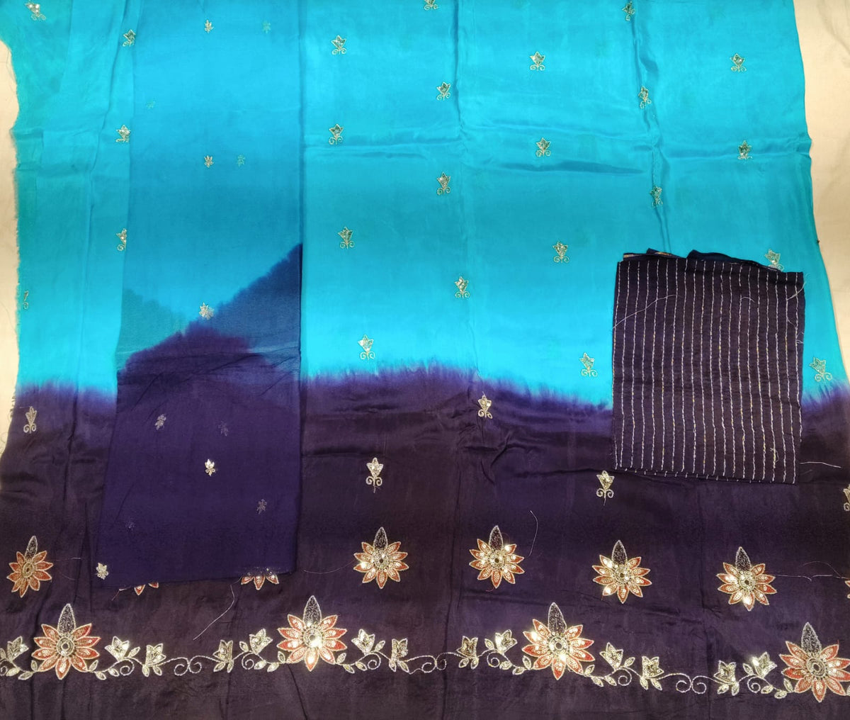 A folded and laid-flat fabric garment, likely a sari or scarf, featuring a vibrant turquoise-to-deep-purple ombré design with metallic gold floral and star motifs, and a striped fabric piece on the right.