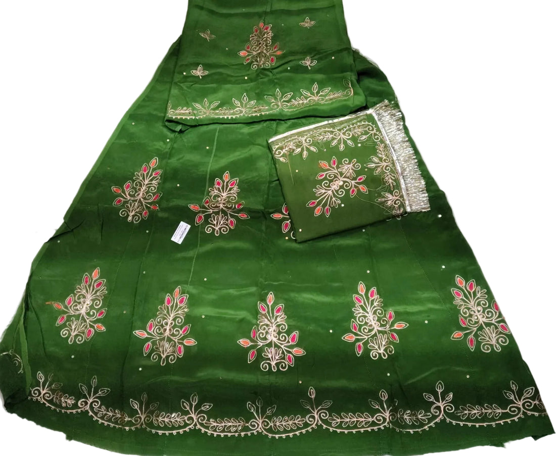 A green traditional fabric set laid flat on a white wrinkled surface, featuring ornate silver and pink floral embroidery with metallic borders. The set includes a skirt, dupatta, and a folded matching fabric piece.