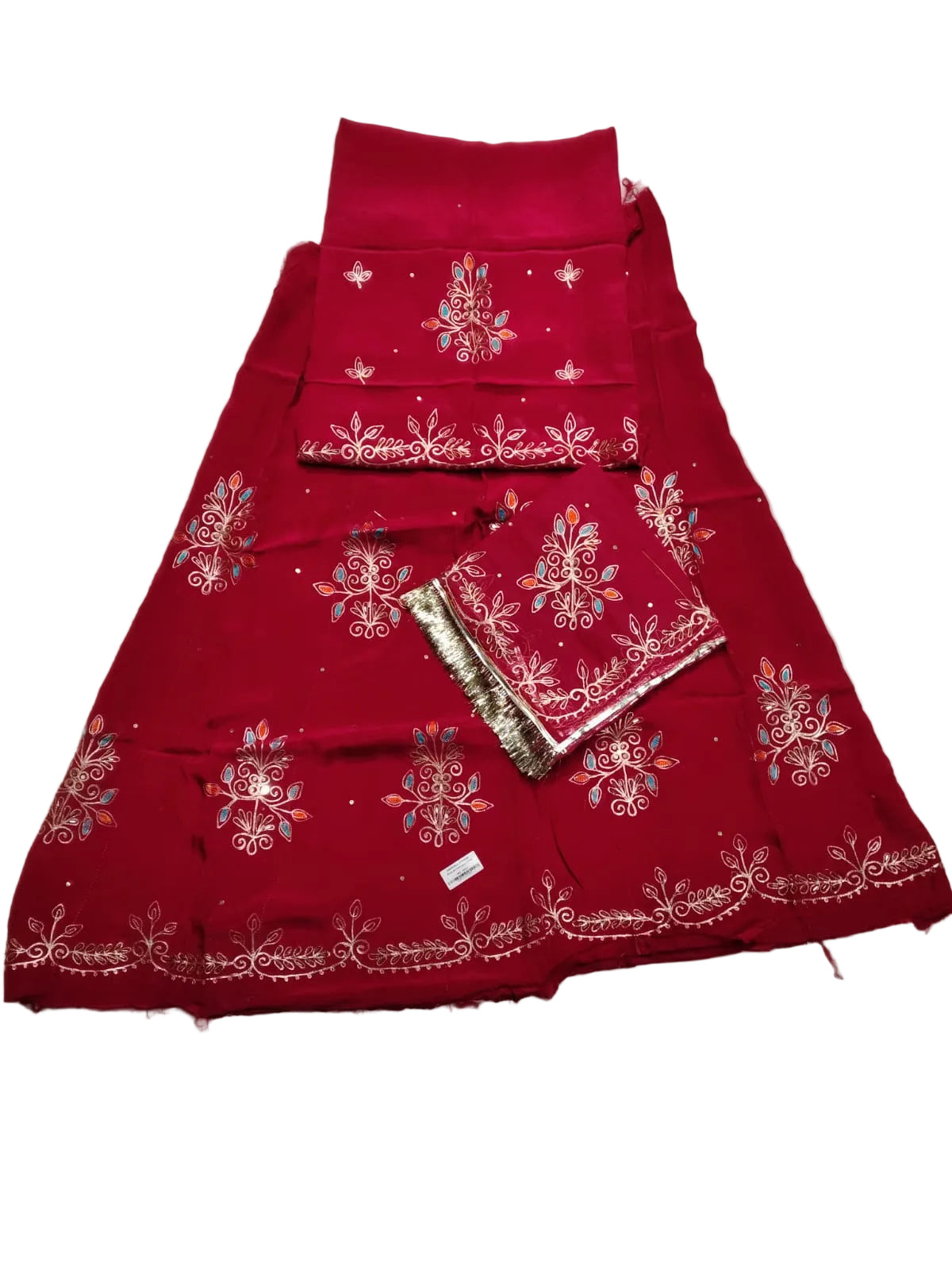 A folded red traditional outfit laid flat on a light gray fabric surface, featuring embroidered floral motifs and metallic border detailing. A matching red dupatta or scarf is placed on top, along with a small metallic accessory.