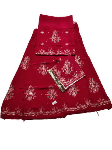 A folded red traditional outfit laid flat on a light gray fabric surface, featuring embroidered floral motifs and metallic border detailing. A matching red dupatta or scarf is placed on top, along with a small metallic accessory.