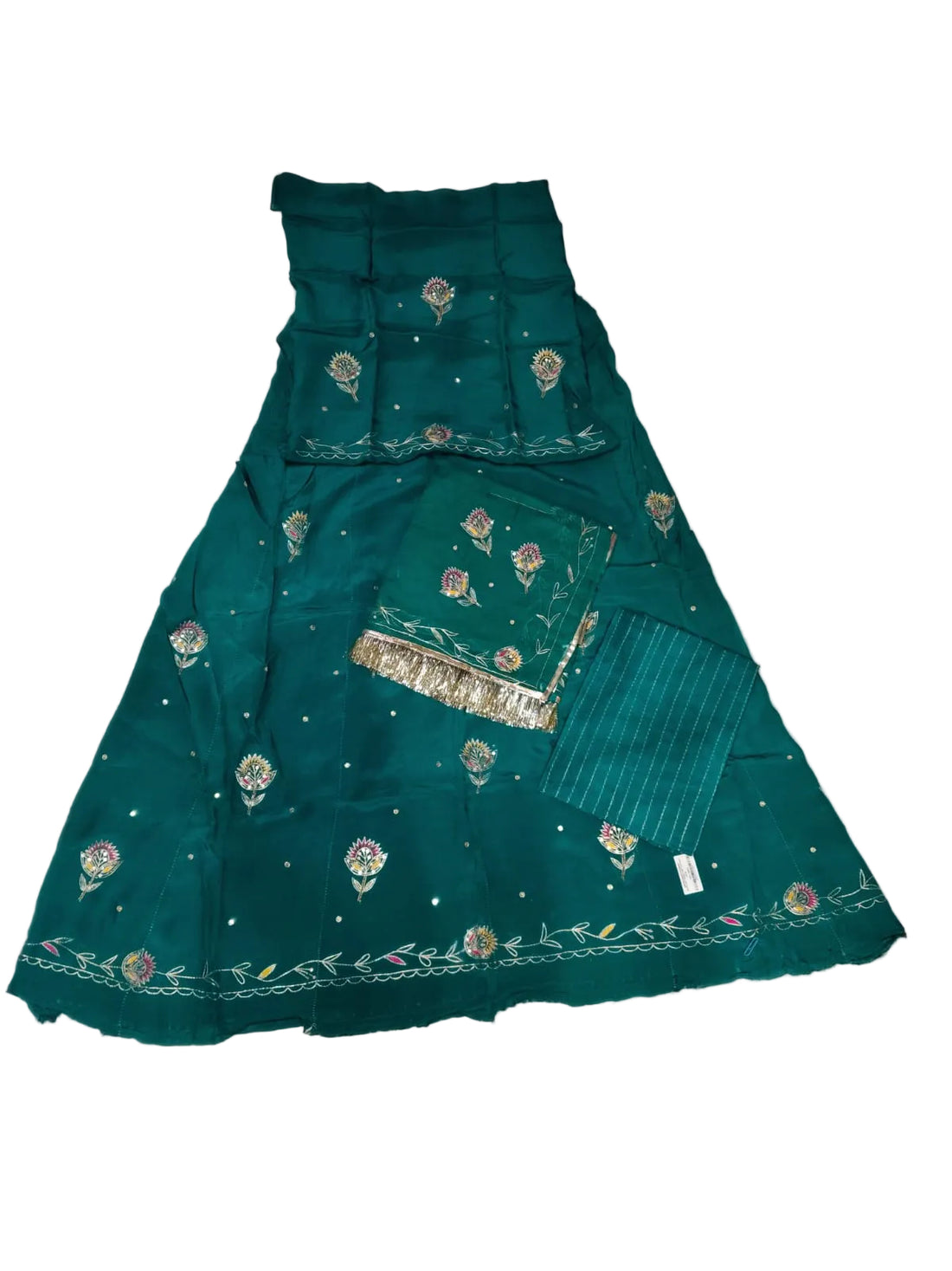 A dark teal green traditional skirt set with floral embroidery and silver mirror work, laid flat on a wrinkled white sheet. The set includes a flared skirt, a matching inner skirt, a striped fabric panel, and a dupatta.