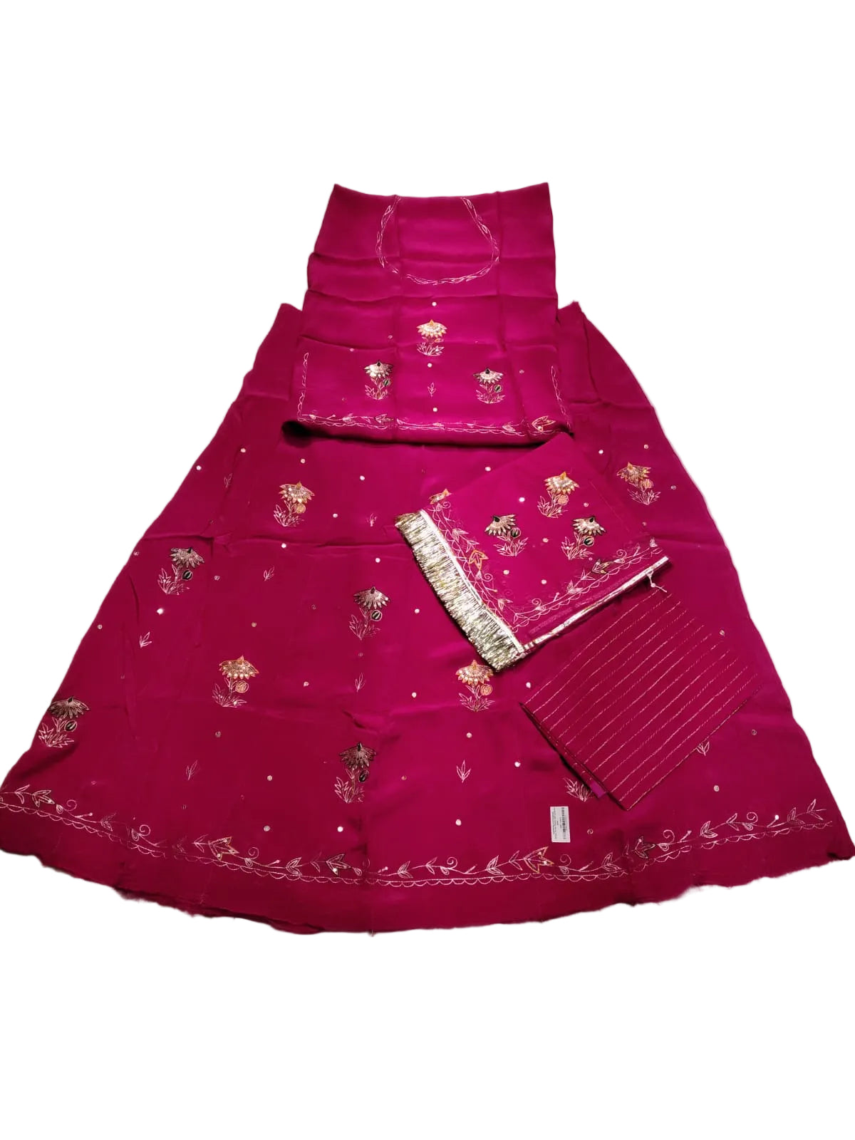 A bright pink traditional Indian outfit set laid flat on a wrinkled light gray sheet. The set includes a flared skirt with silver mirror work and floral embroidery, a matching top, a pleated blouse, and a dupatta.