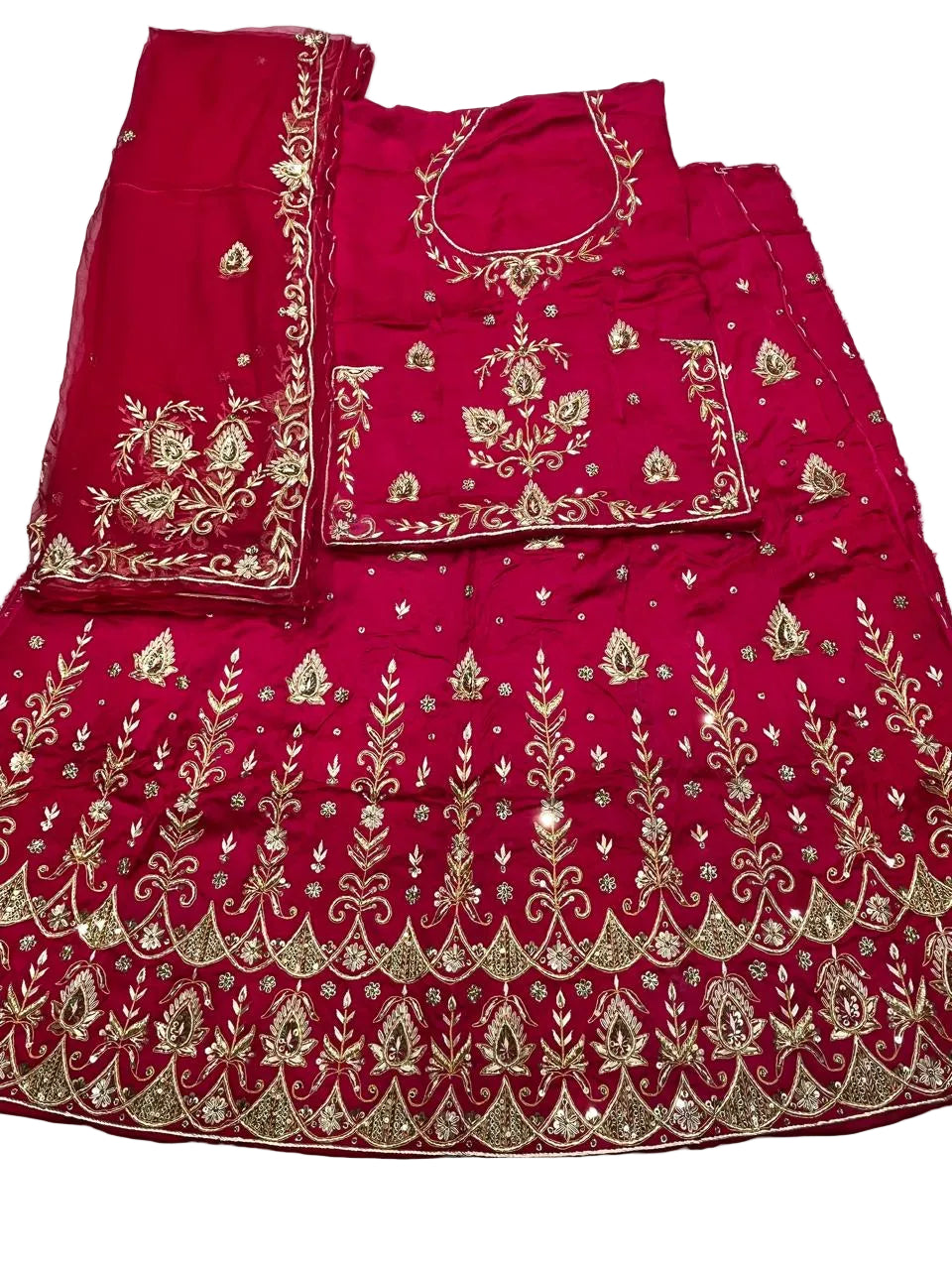 A bright pinkish-red traditional embroidered outfit laid flat on a white fabric surface, including a skirt, matching blouse, and dupatta, decorated with extensive gold metallic embroidery.