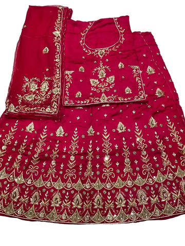 A bright pinkish-red traditional embroidered outfit laid flat on a white fabric surface, including a skirt, matching blouse, and dupatta, decorated with extensive gold metallic embroidery.