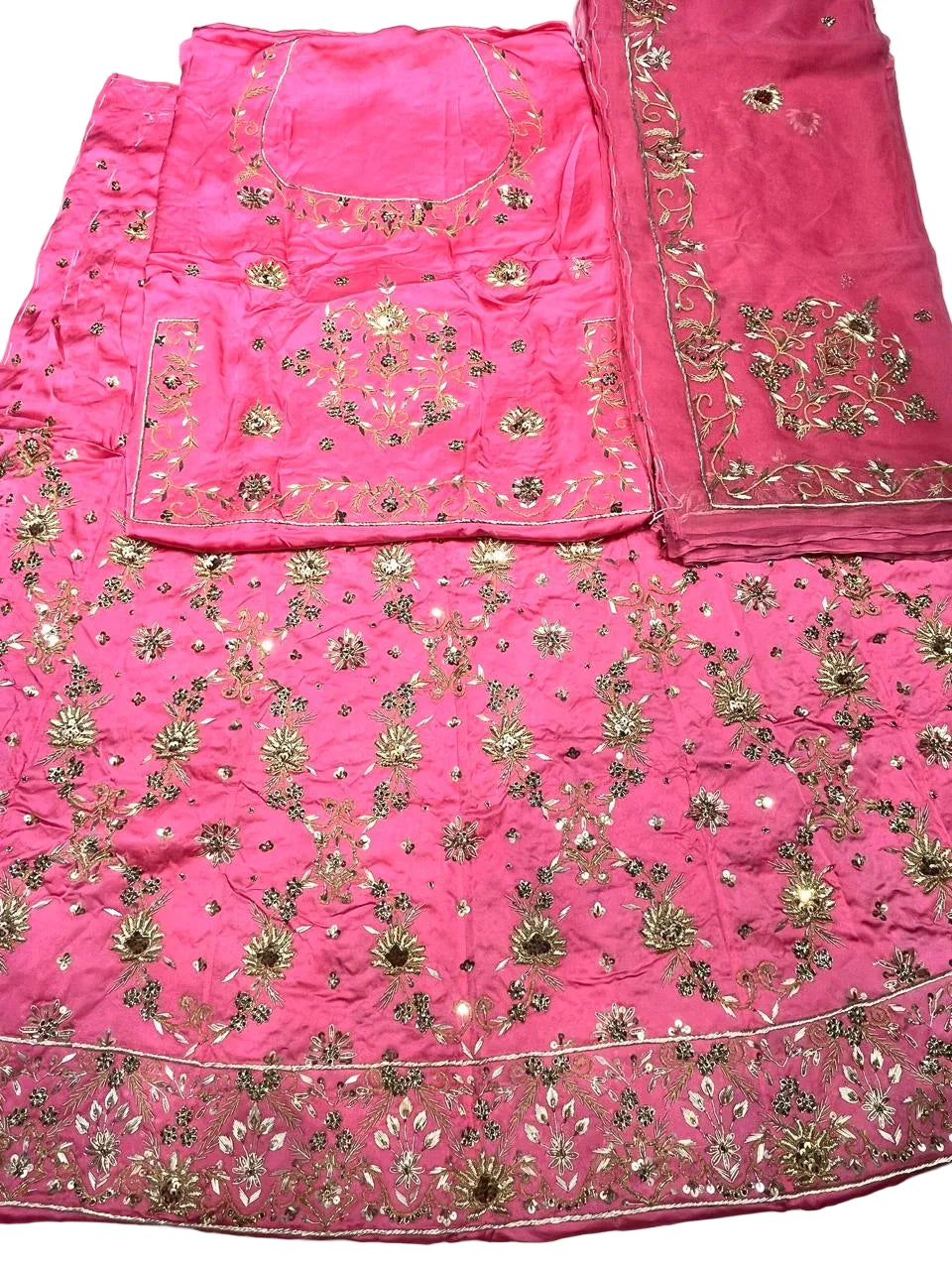 A bright pink traditional embroidered fabric set laid flat on a white surface, including a skirt or drape, matching blouse piece, and dupatta, all decorated with gold floral embroidery and mirror work.