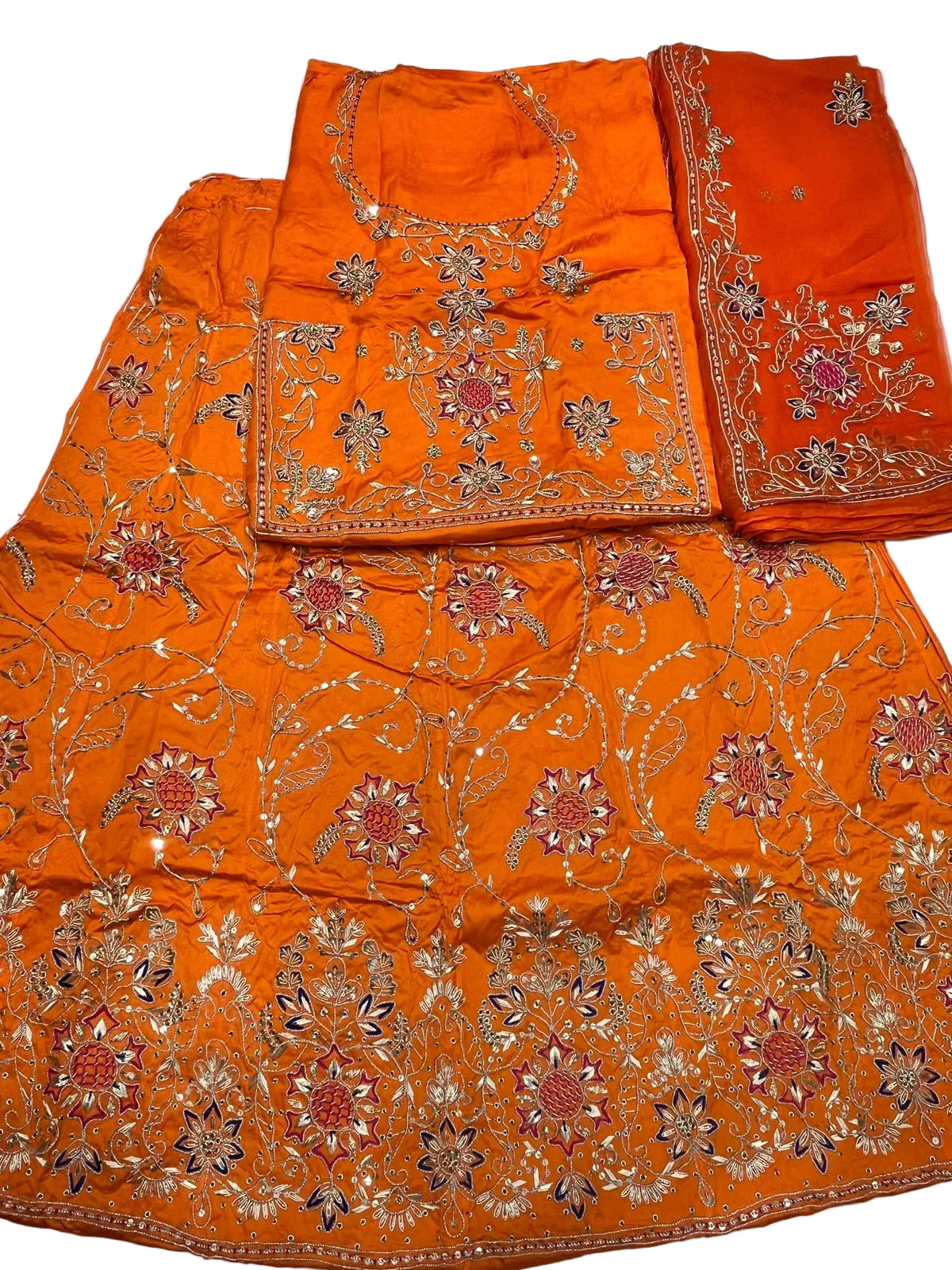 An orange traditional embroidered fabric set laid flat on a white surface, including a skirt or drape, a matching blouse piece, and a folded dupatta or fabric panel with floral embroidery.