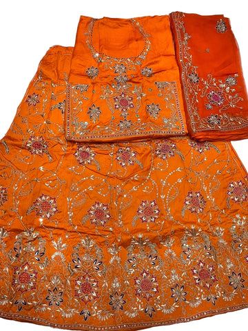 An orange traditional embroidered fabric set laid flat on a white surface, including a skirt or drape, a matching blouse piece, and a folded dupatta or fabric panel with floral embroidery.