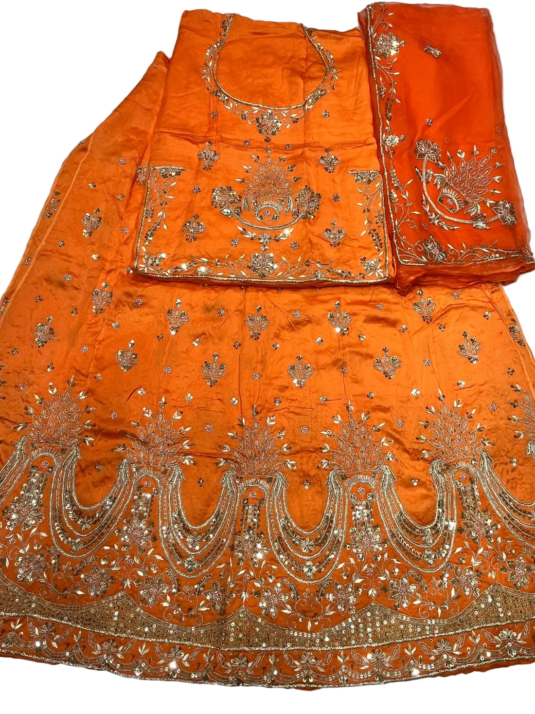 An orange traditional ethnic outfit laid flat on a white fabric background, featuring a kurta, matching skirt, dupatta, and embellished silver mirror work embroidery.